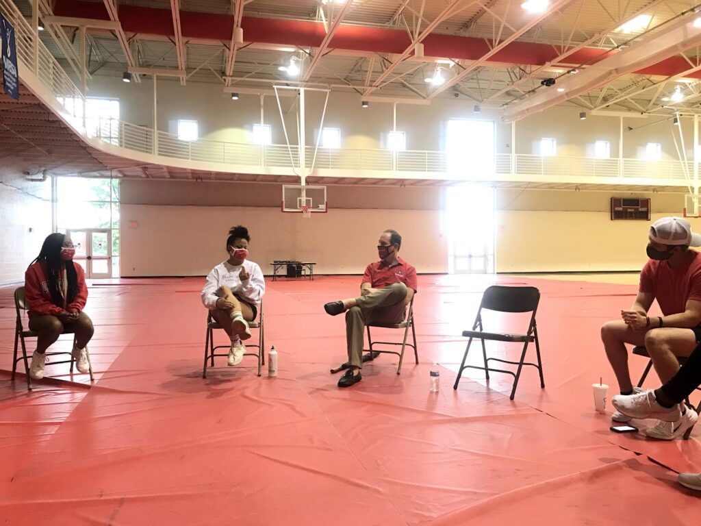 a group sat in chairs separated by two feet in a large circle all wearing masks and trying to talk