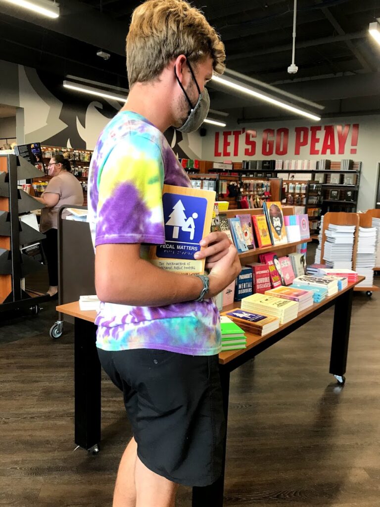 My best friend in the school book store with a mask and a tyedye tshirt on holding a book titled Fecal Matter